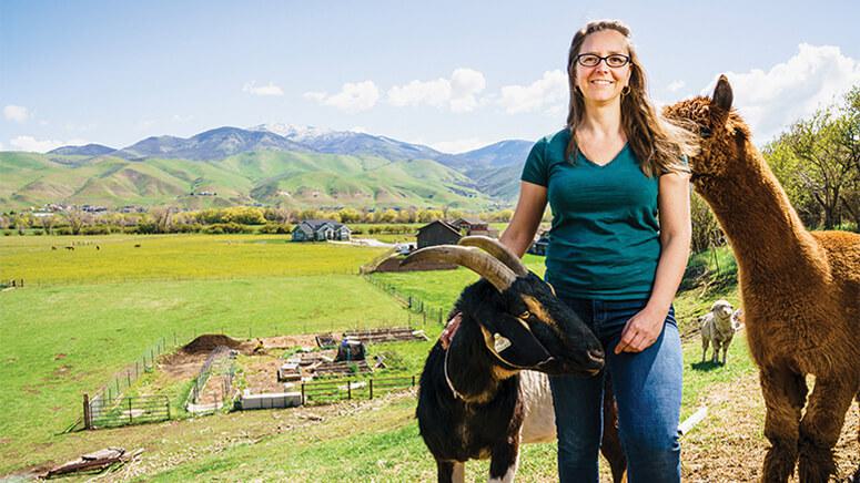 Photo of Sofia Robb standing with a goat and a llama on a farm with mountains in the background.