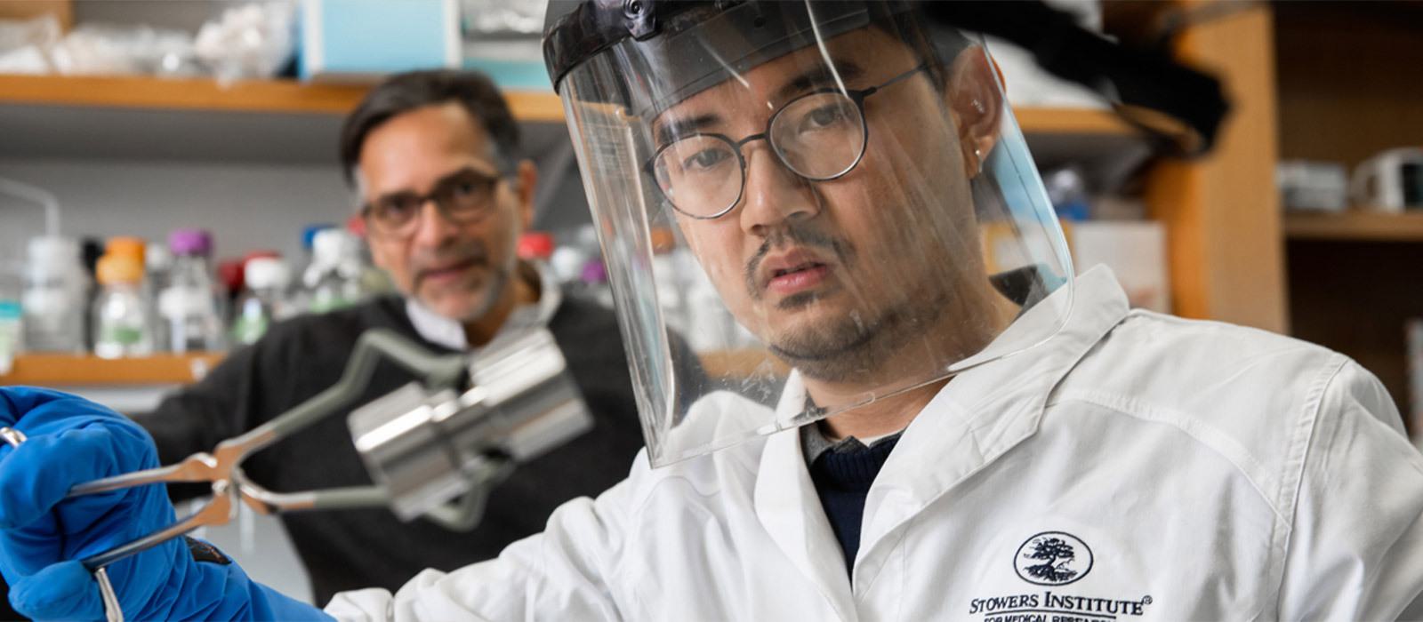 Scientist in lab jacket and face shield holding up metal object