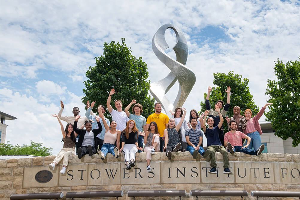 Group of students sitting on Stowers Institute monument sign waving